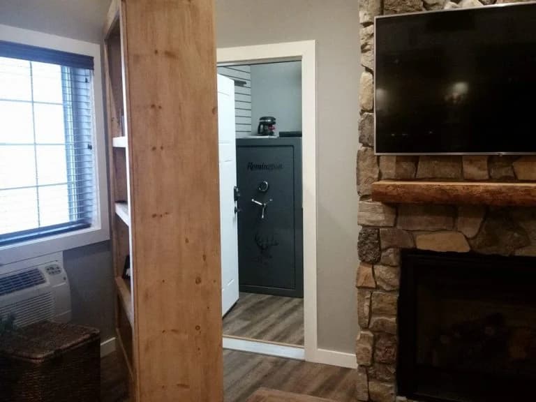 An open concealed doorway beside a stone fireplace reveals a gray safe-room door.